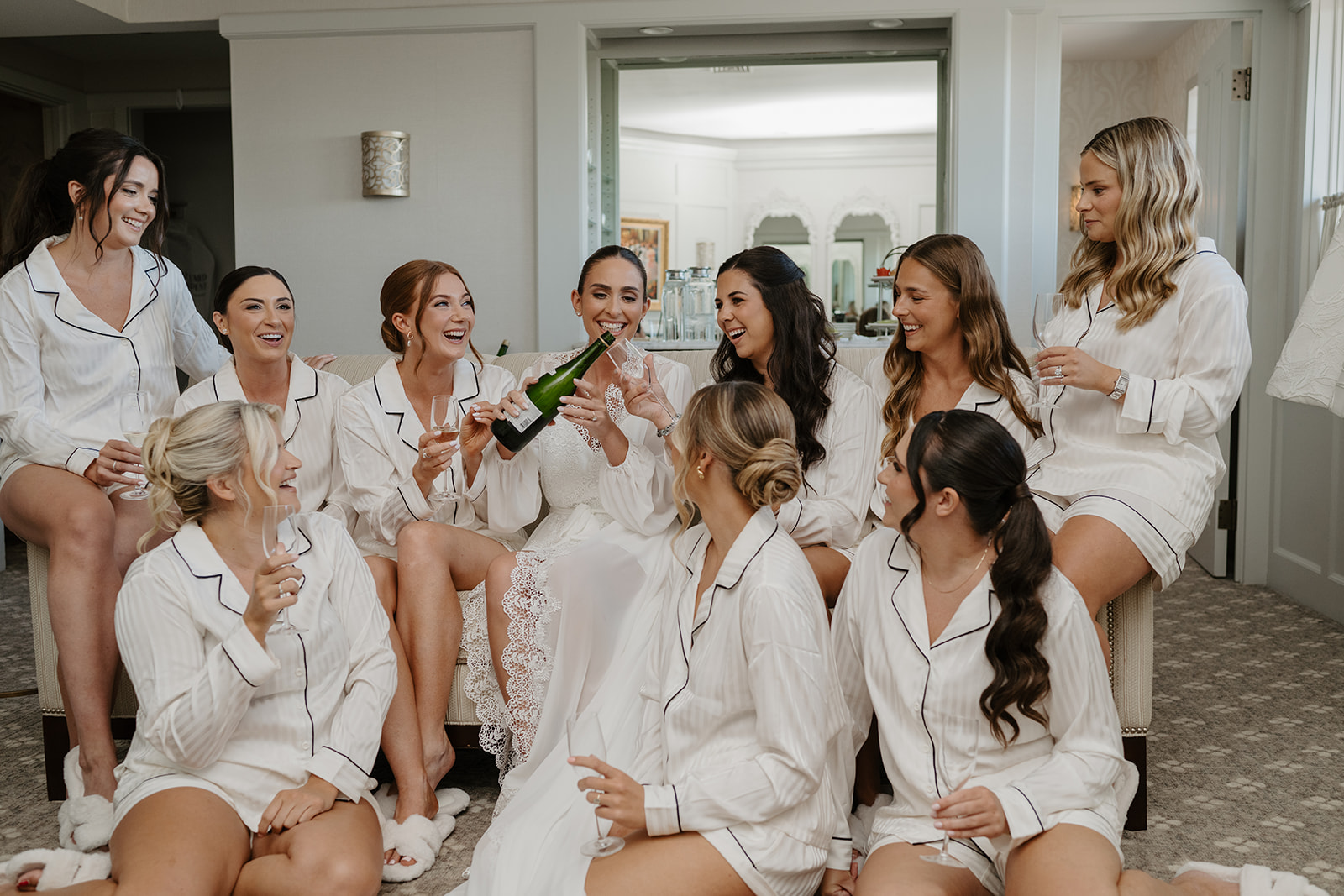 Bride pouring champagne for her bridesmaids in matching pajamas during prep in the bridal suite.